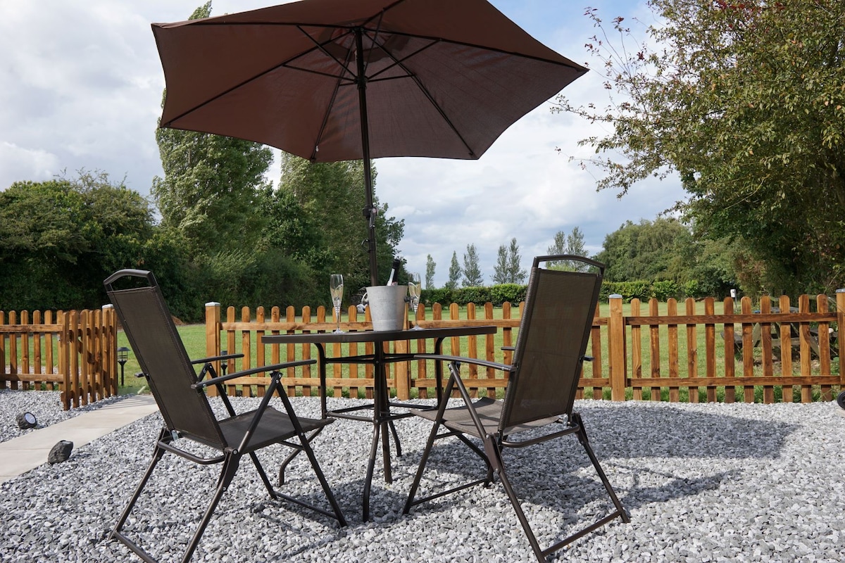 An outdoor patio area features a round table with a large umbrella providing shade. Two folding chairs are positioned around the table, set on a gravel surface. In the background, a wooden fence encloses a green space dotted with trees.