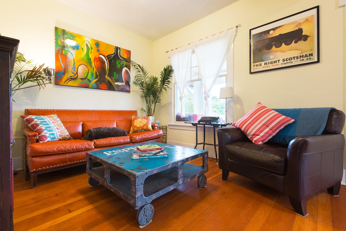 A living room is showcased with hardwood flooring and natural light. A vibrant artwork is displayed above a caramel-colored leather sofa adorned with decorative pillows. A dark brown armchair complements a coffee table with wheels, surrounded by indoor plants near a window.