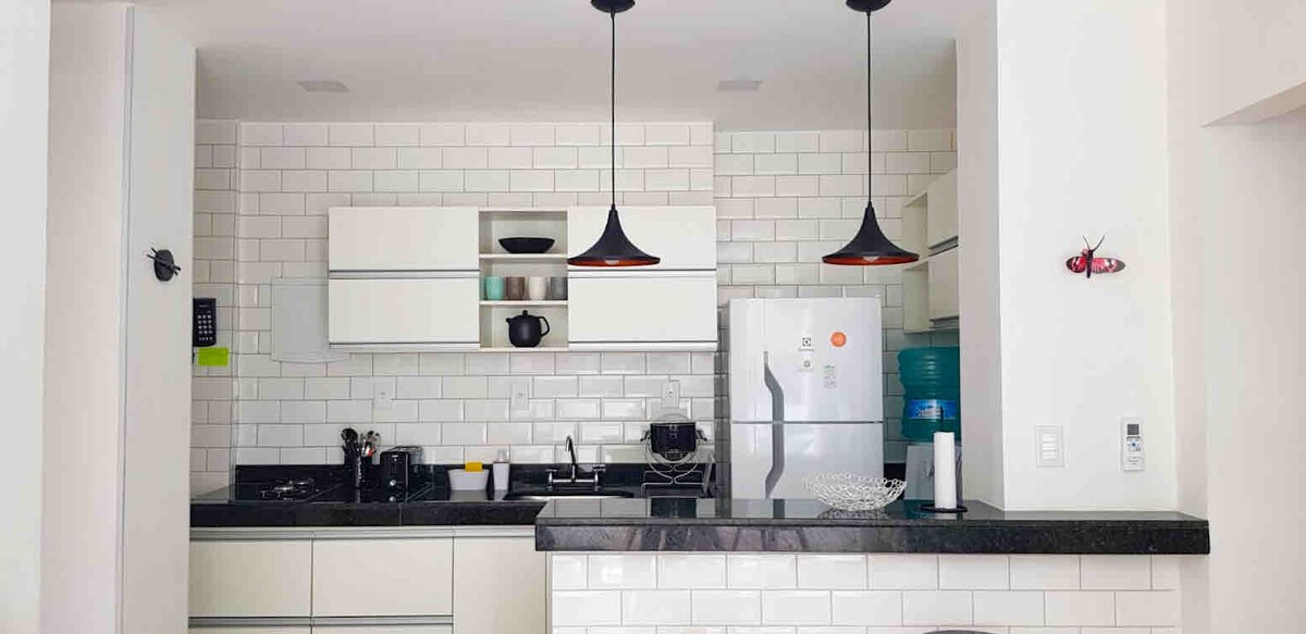 An equipped kitchen features modern cabinetry and sleek black countertops. A refrigerator and water dispenser are visible, along with a gas stovetop and sink. Pendant lights hang above, adding a contemporary touch, while white tiled walls enhance the bright atmosphere.