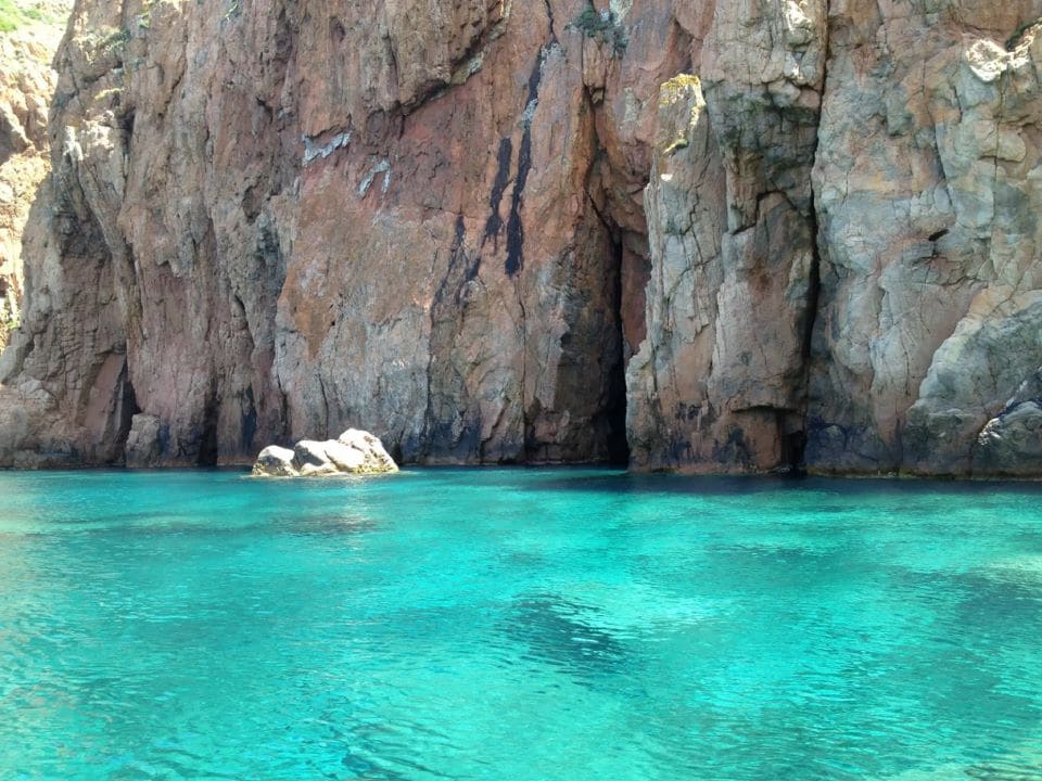 A tranquil turquoise bay is framed by rugged, towering cliffs exhibiting various earthy tones. Smooth, large rocks are partially submerged in the clear water, which reflects light, creating a serene and inviting environment.
