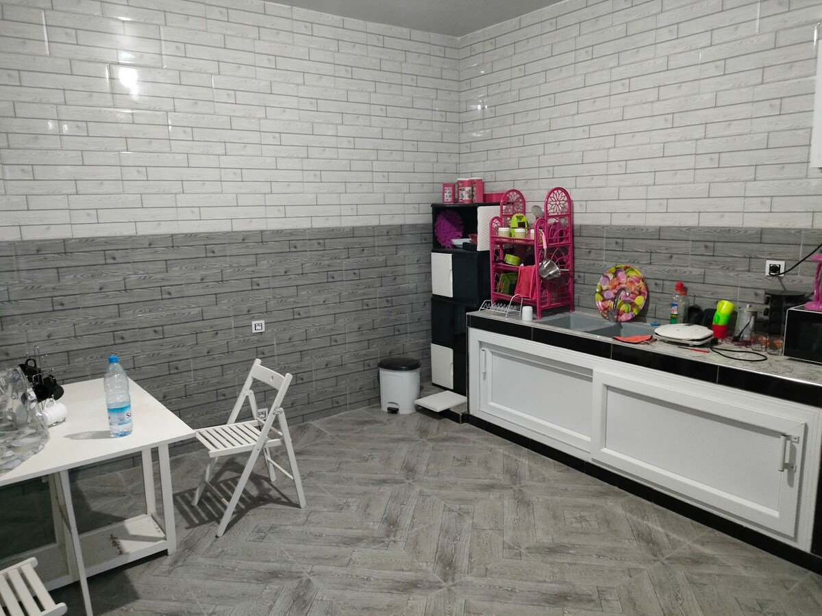 A well-equipped kitchen includes white cabinetry and modern appliances. A small dining table with two folding chairs is positioned near the wall. Colorful dishware and utensils are organized on shelves, while a water bottle is visible on the table.