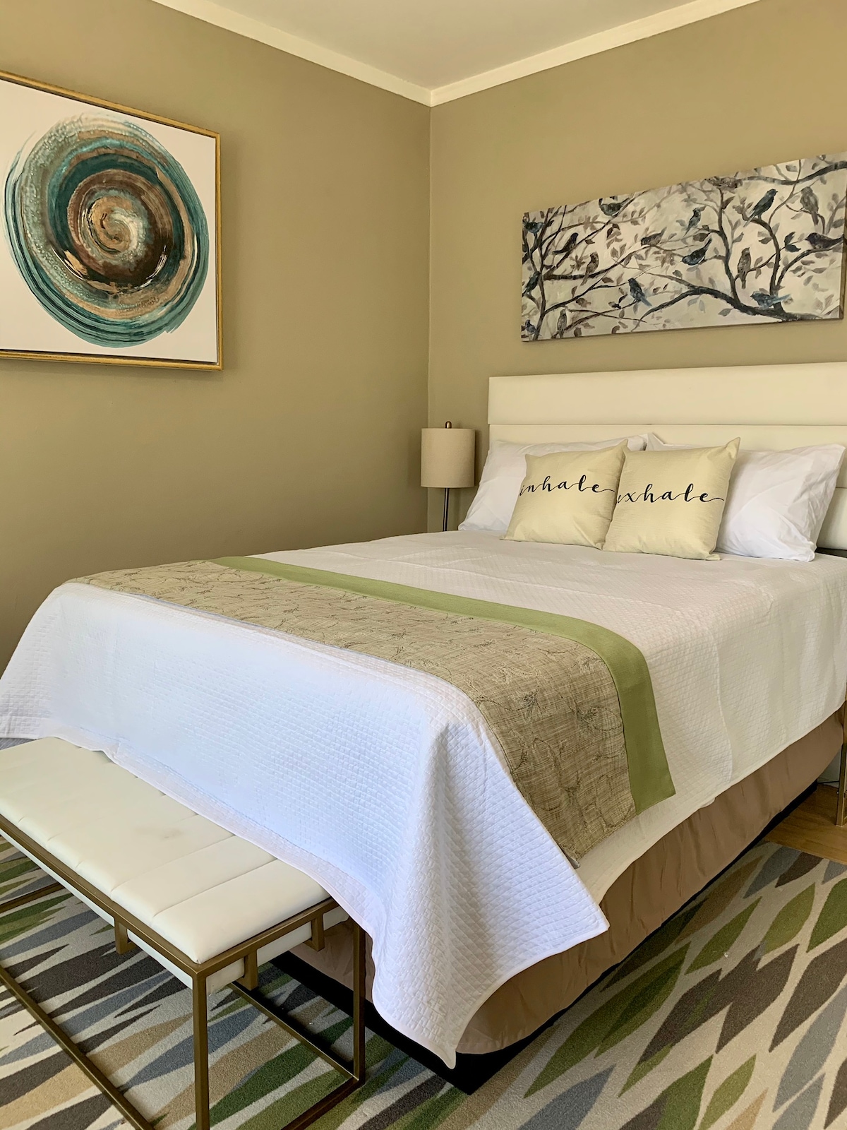 A well-appointed sleeping area features a spacious bed dressed in crisp white linens, accented with decorative pillows. A light-colored bench sits at the foot of the bed. The walls are adorned with abstract art and botanical prints, creating a calming environment.