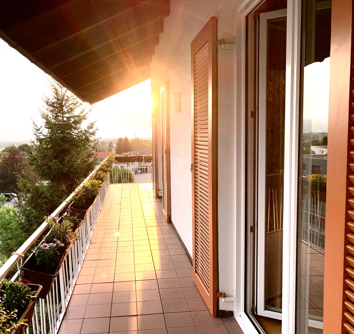 A balcony is presented with a tiled floor and surrounded by flower planters. Doors leading into the interior are partially opened, allowing natural light to enhance the space. The glow of sunset creates a warm atmosphere while offering a view of the surrounding landscape.