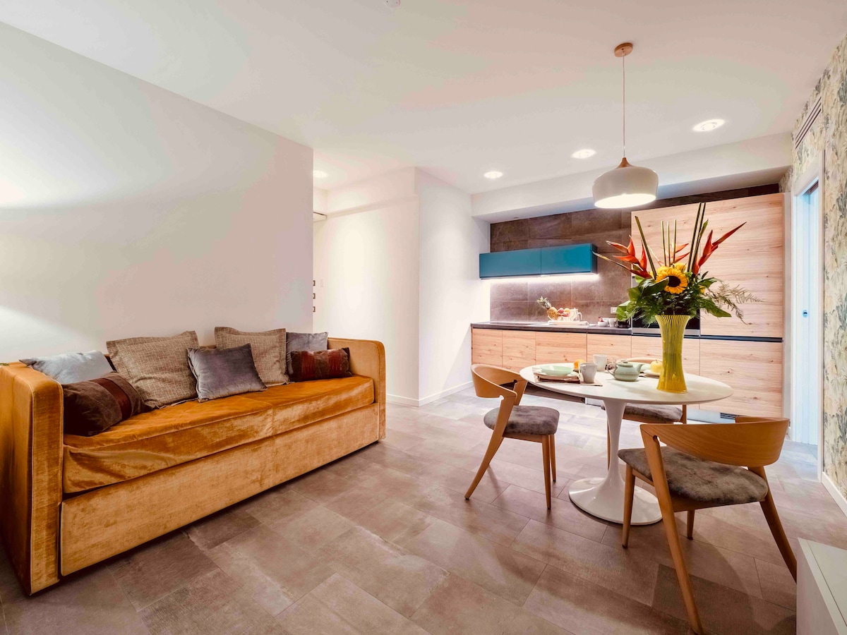 A well-lit living space is shown, featuring a plush sofa adorned with decorative pillows. A round dining table with chairs is positioned near a modern kitchen, highlighted by wooden cabinetry and vibrant floral arrangements on the table.