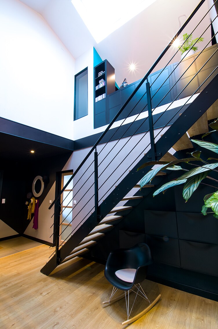 An open and contemporary entryway features a sleek staircase with wooden steps and a metal railing. A large skylight allows natural light to fill the space. A black rocking chair is positioned against a textured wall, adding a modern touch to the design.