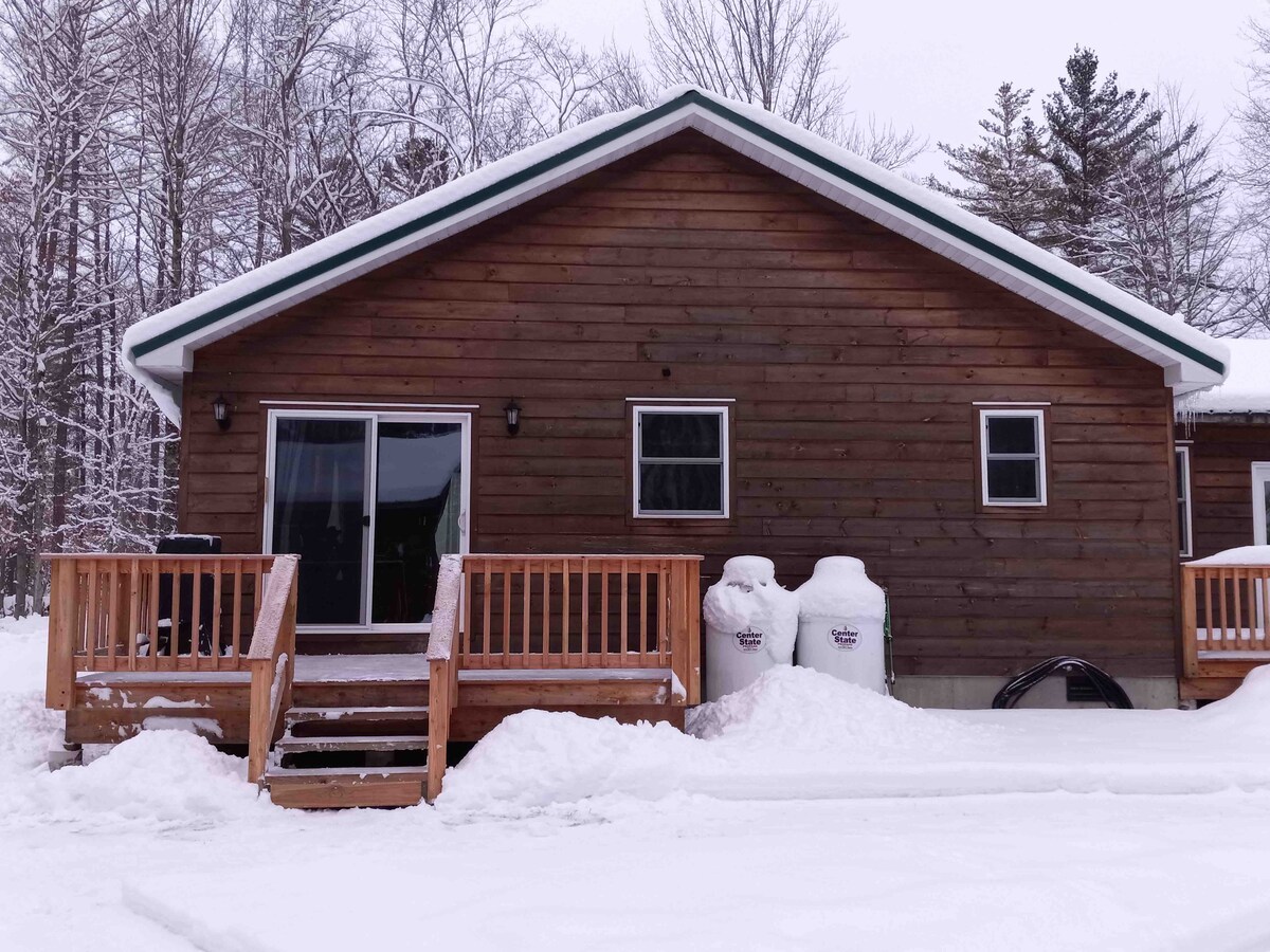 The exterior of a rustic wooden cabin is shown, featuring a sloped roof and large glass doors leading to a porch. Snow covers the ground, and trees surround the property, providing a serene and natural backdrop.