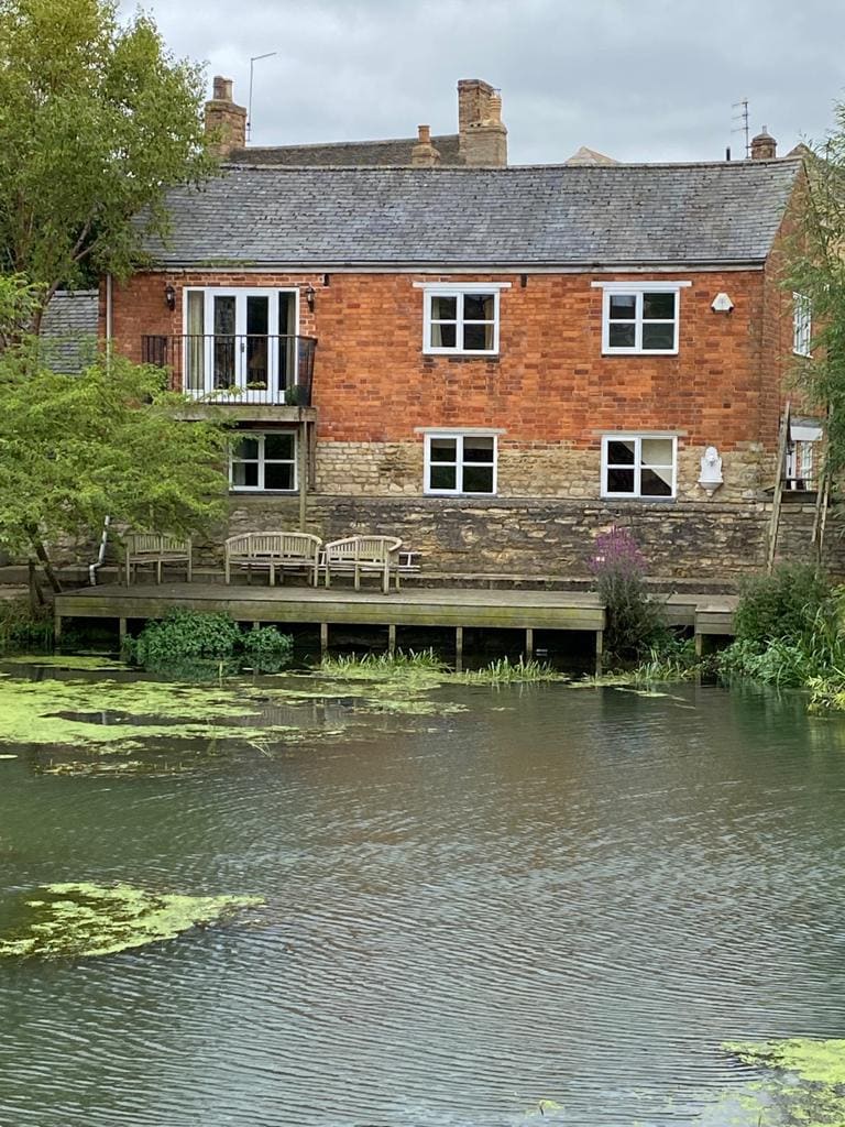 The townhouse features a rustic brick exterior, complemented by multiple windows and a balcony overlooking the river. A wooden deck with seating areas extends over the water, surrounded by greenery and water plants.