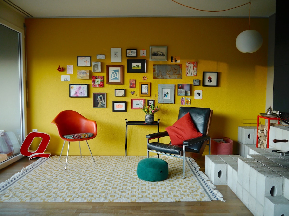 A bright living area features a vibrant yellow wall adorned with an array of framed artworks and photographs. A red chair with a floral seat sits beside a small black table, which holds a vase with fresh flowers. A cozy black armchair and a green ottoman complete the space.