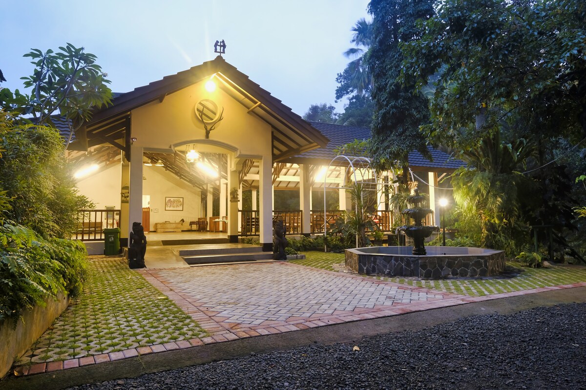 The villa's entrance is framed by lush greenery and stone pathways, leading to a welcoming front porch. Soft outdoor lighting accentuates the structure's architectural features, while a water fountain adds a serene touch to the inviting atmosphere.