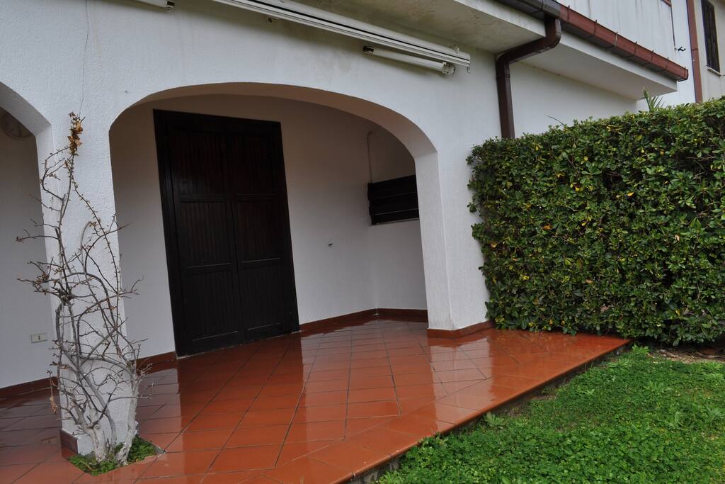 Ingresso alla villa con porticato, giardino, vialetto e doccia esterna.