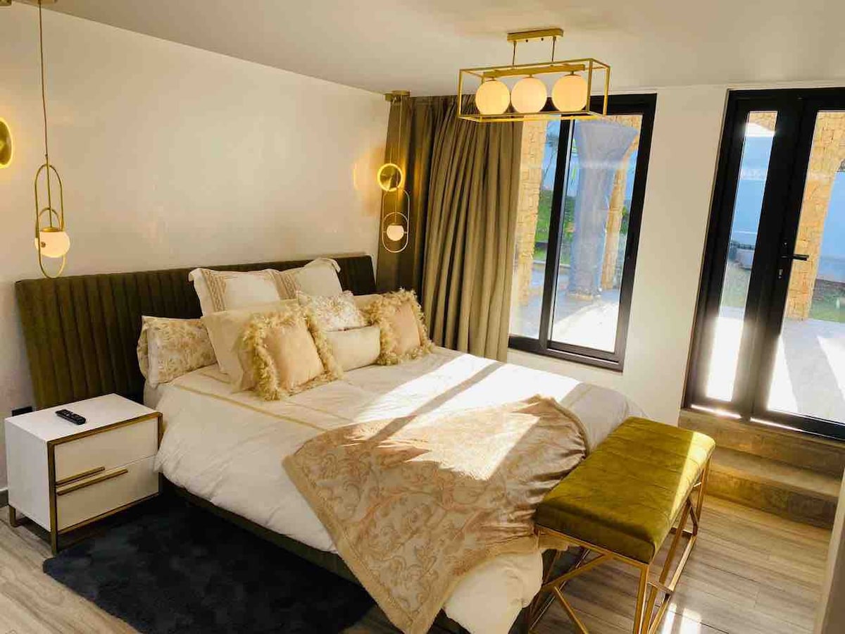 The sleeping area features a spacious bed adorned with decorative pillows and a soft throw blanket. Modern lighting fixtures provide illumination, while large windows allow natural light to fill the room. A stylish bench complements the bed, creating a cozy and welcoming ambiance.