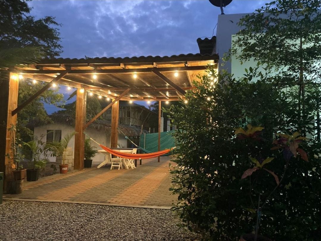 A covered outdoor area is illuminated by warm string lights, providing a cozy ambiance. A vibrant hammock hangs between wooden posts. Nearby, a small table with chairs is positioned on a paved surface surrounded by lush greenery and plants.