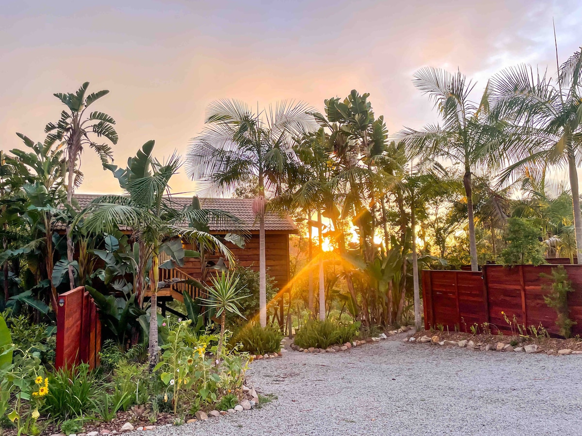 A stilted wooden chalet is surrounded by lush greenery and palm trees. The gravel path leads to the entrance, where a warm glow from the setting sun filters through the leaves, creating a serene atmosphere. Fenced areas and landscaping add to the natural beauty.