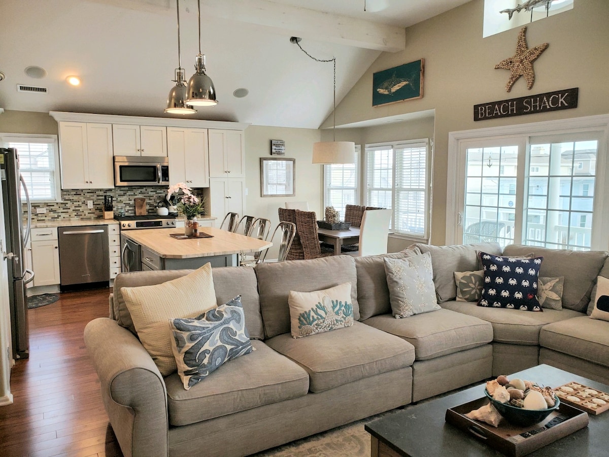 An open concept living space features a comfortable gray sectional sofa adorned with patterned throw pillows. A light wood table accommodates several chairs, while a fully equipped kitchen showcases stainless steel appliances and a large island. Natural light floods in through multiple windows.