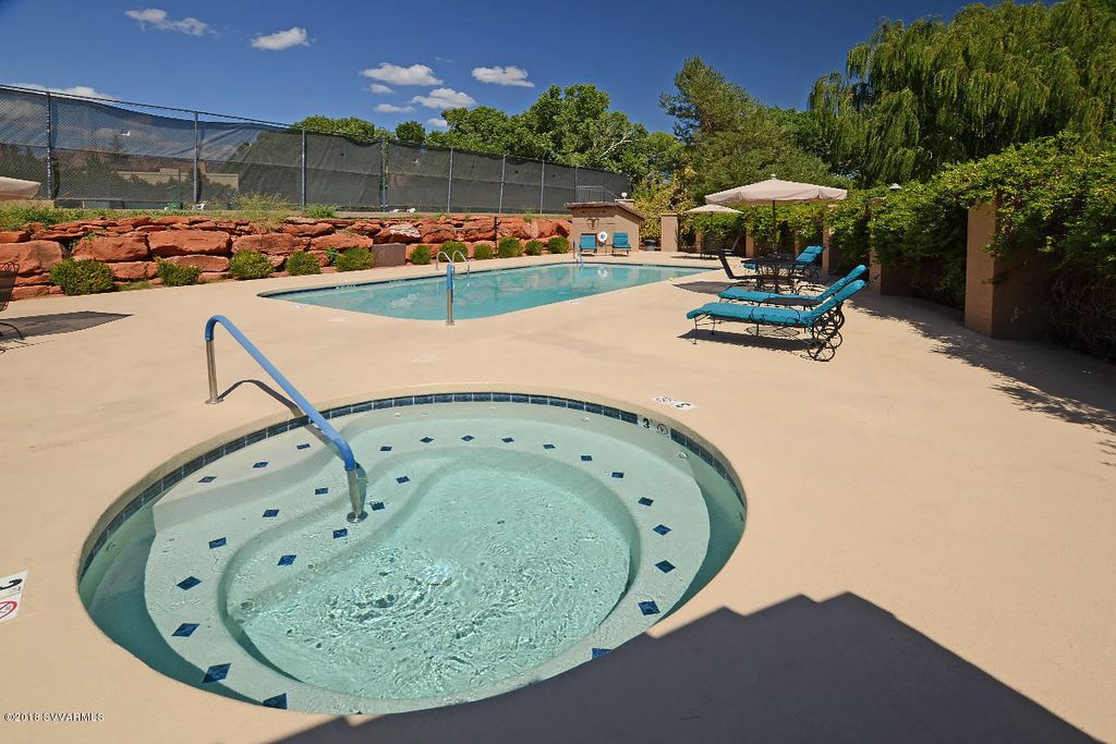 A hot tub and a spacious pool are positioned on a concrete patio, surrounded by vibrant greenery and stone landscaping. Lounge chairs sit under shaded umbrellas, creating a relaxing outdoor space. A clear blue sky is visible overhead, enhancing the inviting atmosphere.