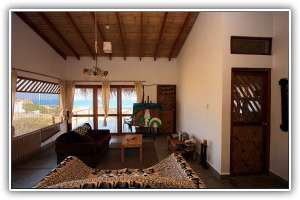 An airy living space with a high wooden ceiling is showcased, featuring large windows that offer views of the sea. A dark sofa and a wooden table provide seating, while the walls are partially adorned with decorative accents. Light filters through a nearby window, enhancing the serene atmosphere.