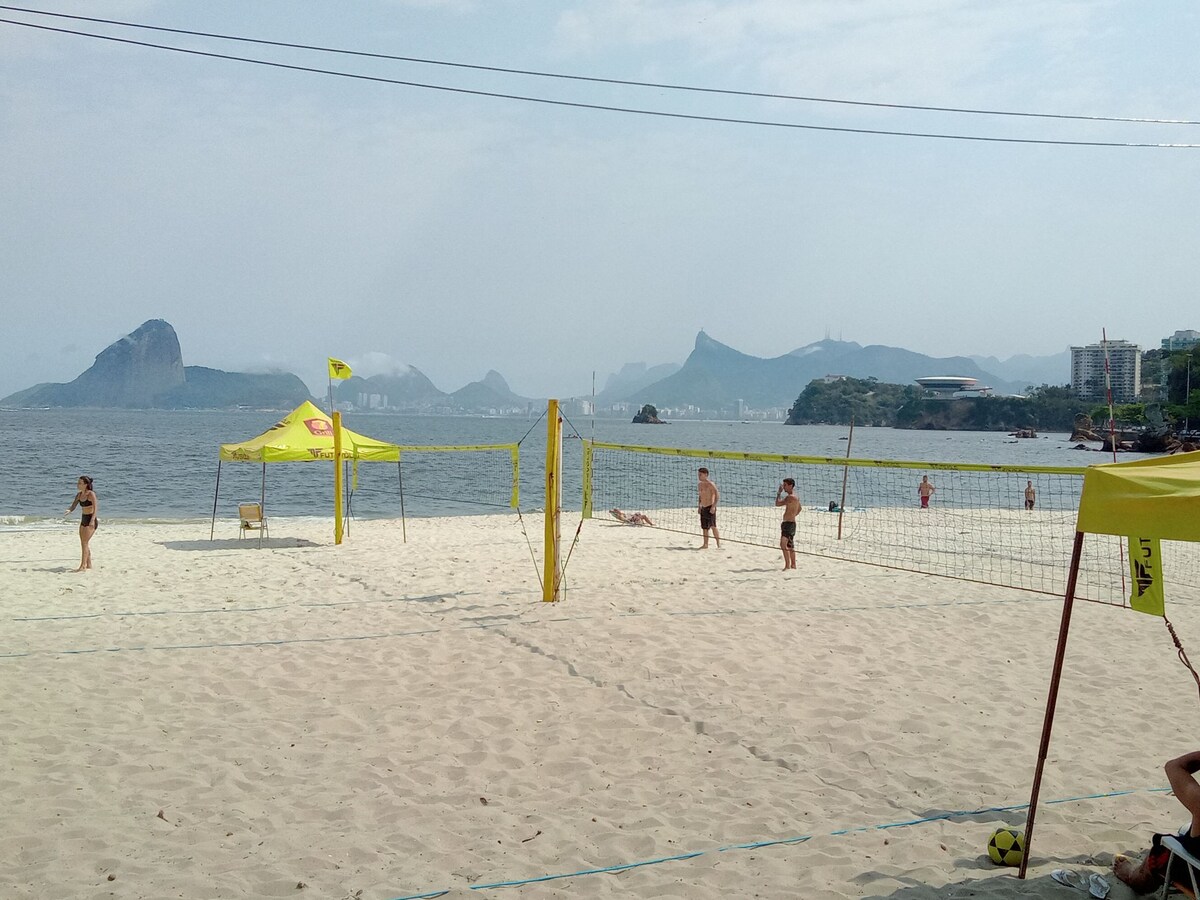 A sandy beach scene is depicted, featuring beach volleyball courts with players engaged in a game. The ocean stretches across the horizon, and mountains are visible in the distance under a cloudy sky. Bright yellow tents provide shaded areas along the beach.