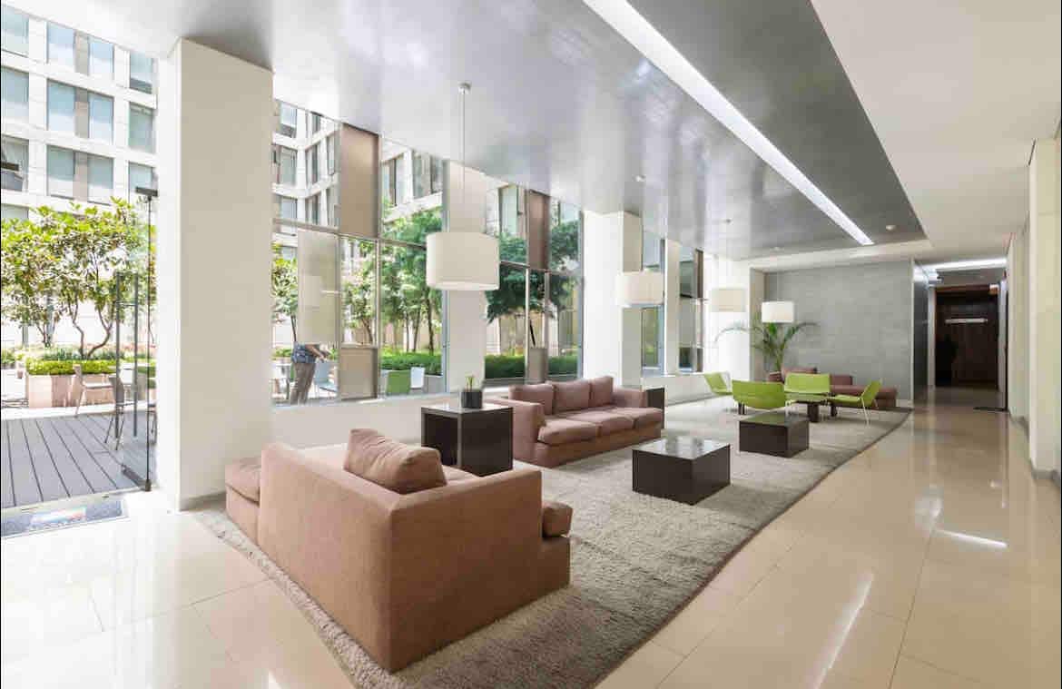 A spacious lobby area features large windows allowing natural light to illuminate the seating arrangements. Soft-colored sofas and green armchairs are arranged around low, dark tables, complemented by modern light fixtures hanging from the ceiling.