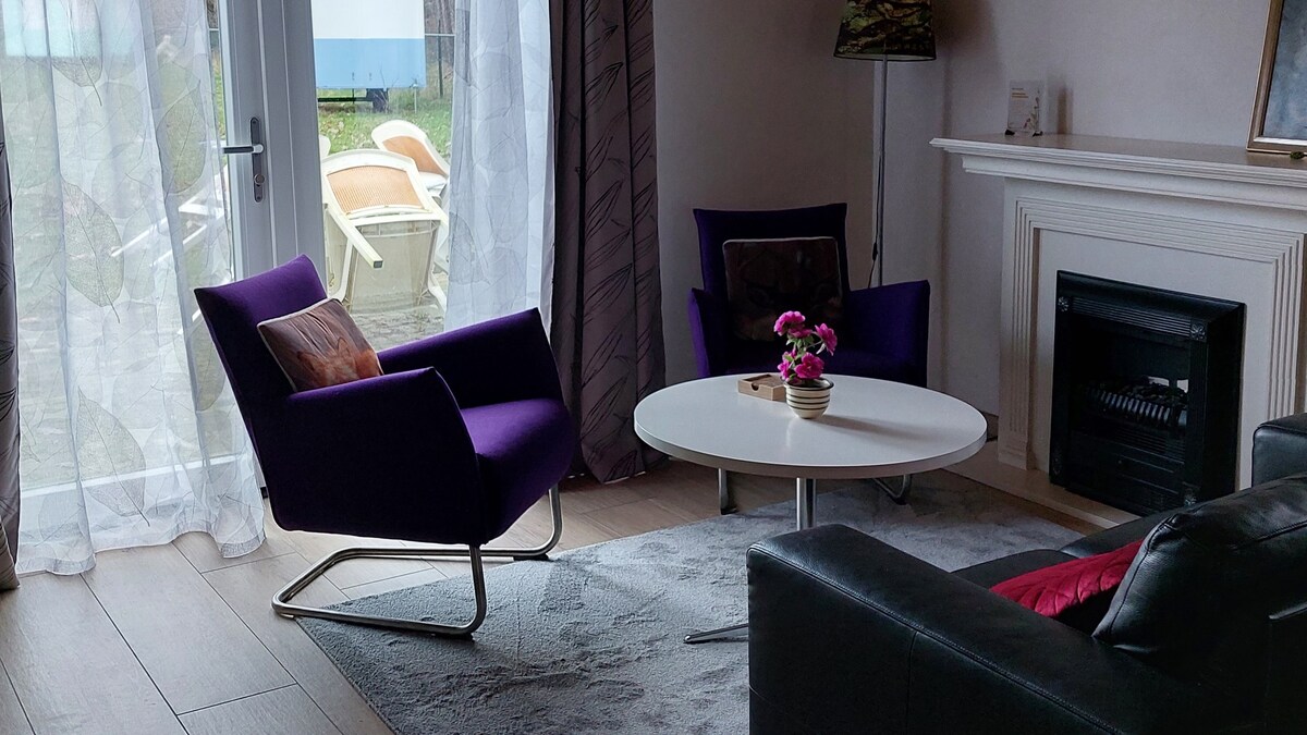 A living area is showcased with two purple chairs positioned near a modern coffee table. A decorative plant sits in the center of the table. Natural light filters through sheer curtains, illuminating the space beside a cozy fireplace.