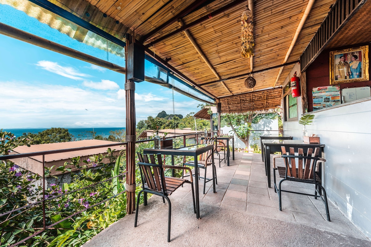 An open terrace features multiple wooden tables and chairs, offering unobstructed views of the ocean and surrounding greenery. The roof is constructed with bamboo, providing shade and a rustic charm. Potted plants add a touch of nature, enhancing the inviting atmosphere.