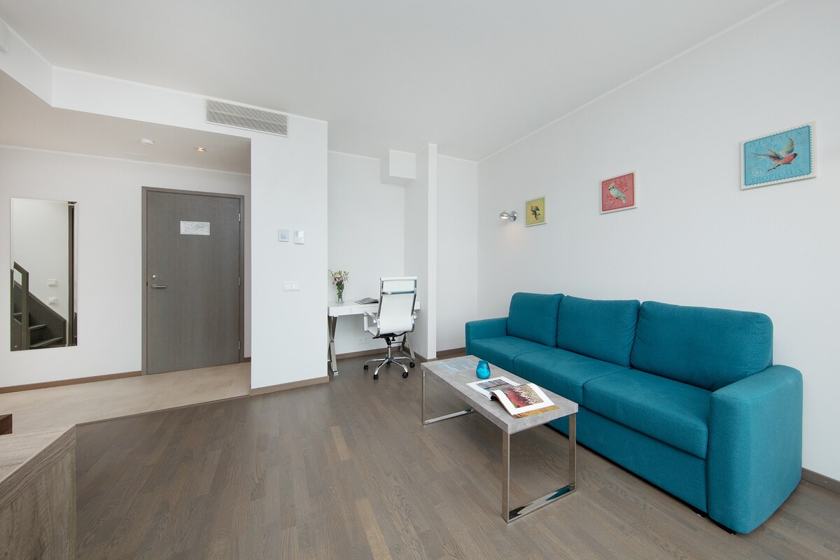 The living area features a spacious teal sofa positioned across from a modern coffee table. A white desk with a chair is nearby, complemented by artwork on the walls. The entrance door is visible, along with a mirror reflecting part of the room.