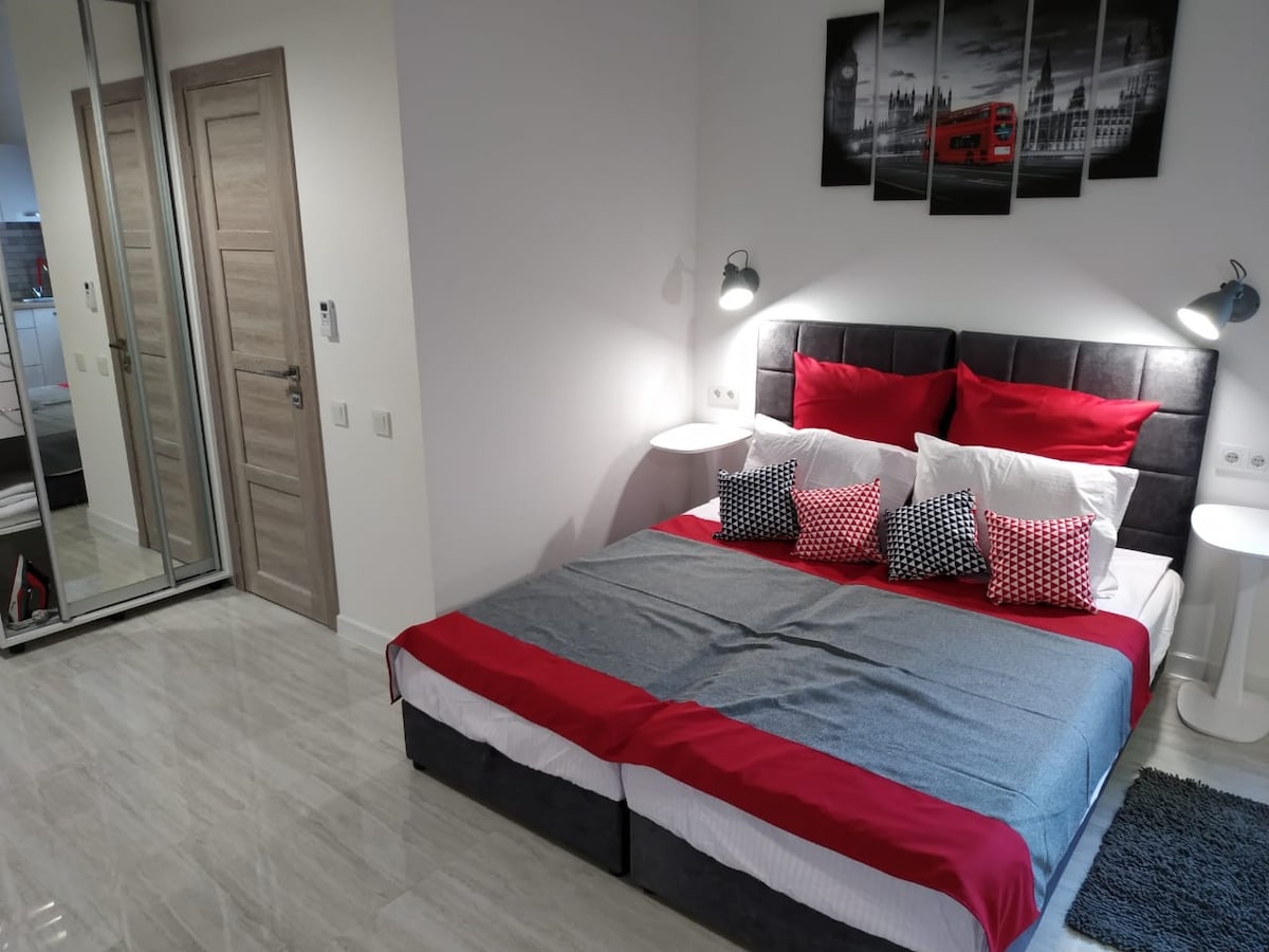 A cozy sleeping area features a spacious bed with red and gray linens, accented by patterned pillows. Two bedside tables are positioned on either side, and a mirror reflects the inviting ambiance. Soft light is provided by wall-mounted lamps.