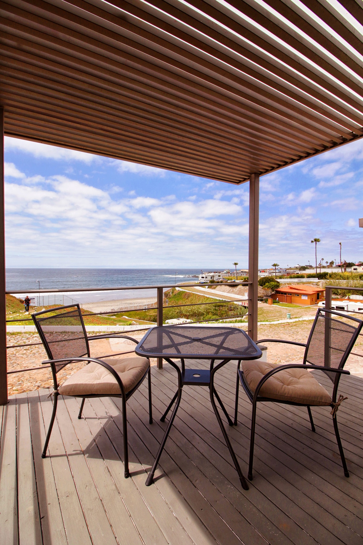 An outdoor patio features a small black table with two beige chairs, framed by a railing overlooking the ocean. The sky above is partly cloudy, casting soft light on the tranquil water, while the shore can be seen in the distance.