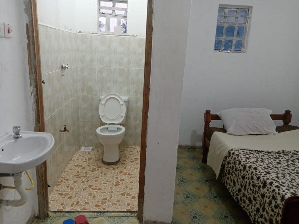 An interior space includes a simple bedroom with a bed and light-colored blanket, alongside a bathroom area featuring a tiled floor and a shower with a toilet. Natural light enters through a small window, illuminating the neutral-toned walls.