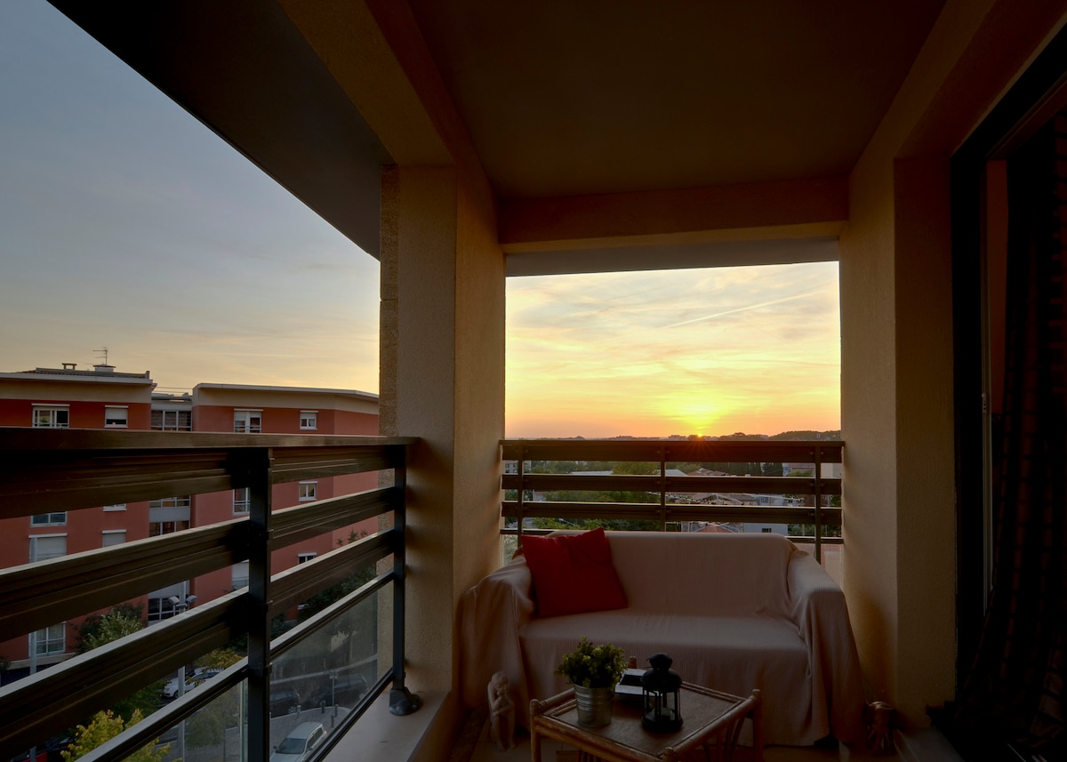 A terrace features a comfortable seating area with a cozy blanket draped over the sofa. A small table holds decorative items. The sunset is visible through large windows, casting warm hues across the space, while the neighboring buildings outline the horizon.