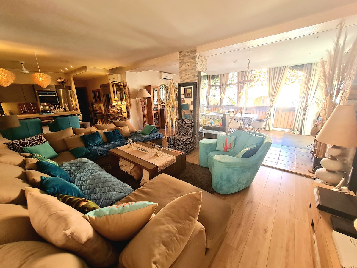 A spacious living area features a large sectional sofa adorned with colorful cushions. A wooden coffee table sits at the center, surrounded by two distinct armchairs. Sliding glass doors lead to an outdoor terrace, allowing natural light to fill the room.