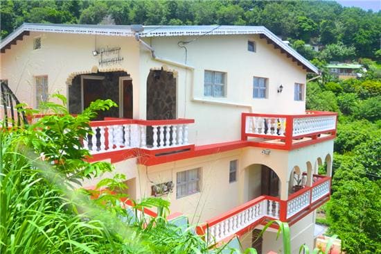 A three-tiered structure is surrounded by lush greenery, showcasing a combination of balconies and terraces with white railings. The exterior features light-colored walls and stone accents, complemented by red trim. The tranquil hillside location offers a serene atmosphere against a backdrop of trees.