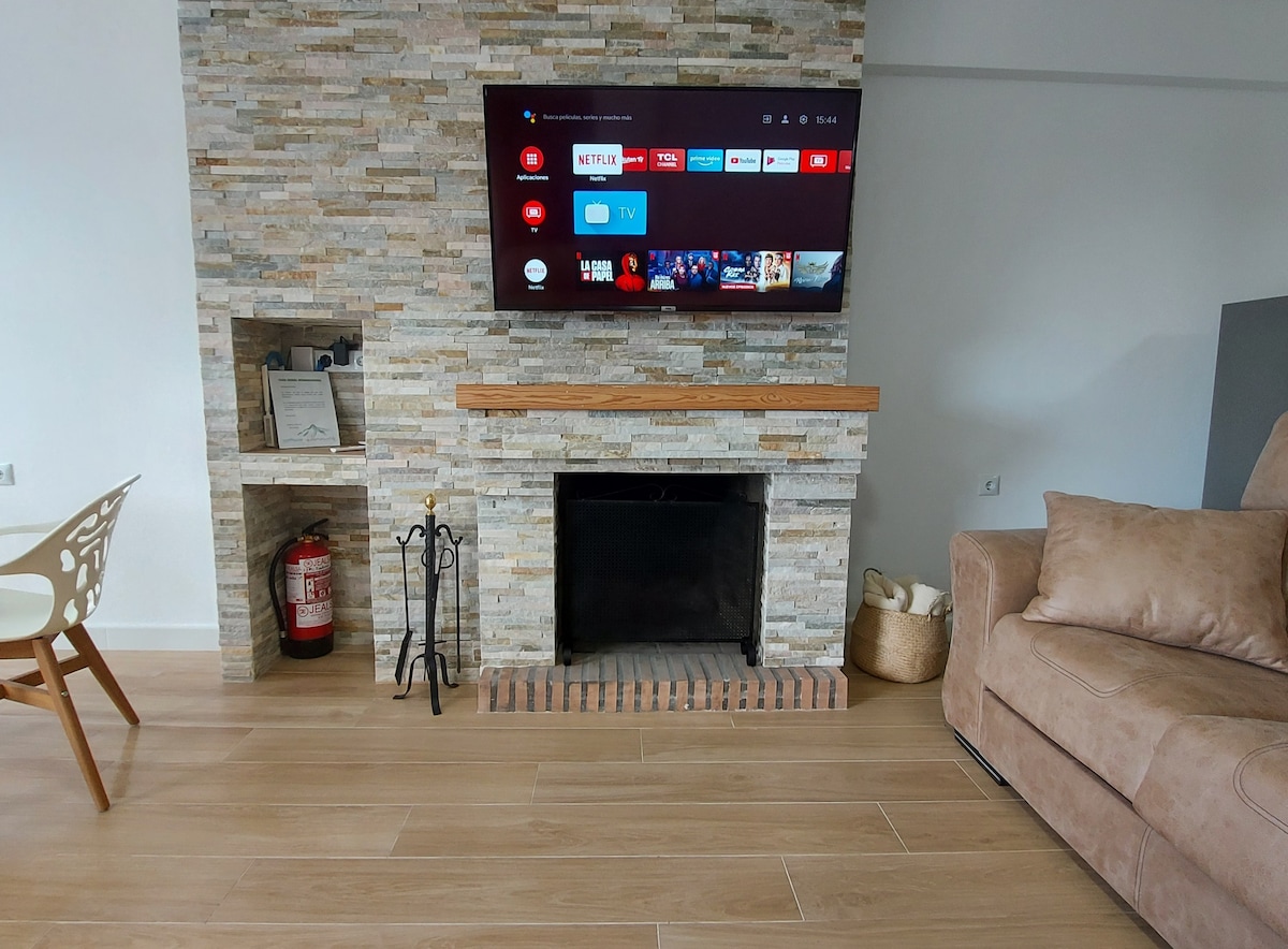 A modern living area features a stone fireplace with a flat-screen TV mounted above. A comfortable sofa sits nearby, and a dining table with stylish chairs is visible. Floor tiles in a warm wood tone enhance the inviting ambiance of the space.