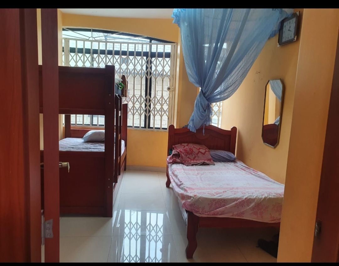 A cozy sleeping area is equipped with two wooden beds, one larger and one bunk-style. The walls are painted a warm yellow, and light enters through a window protected by a security grille. Soft bedding and a decorative curtain add a touch of comfort.