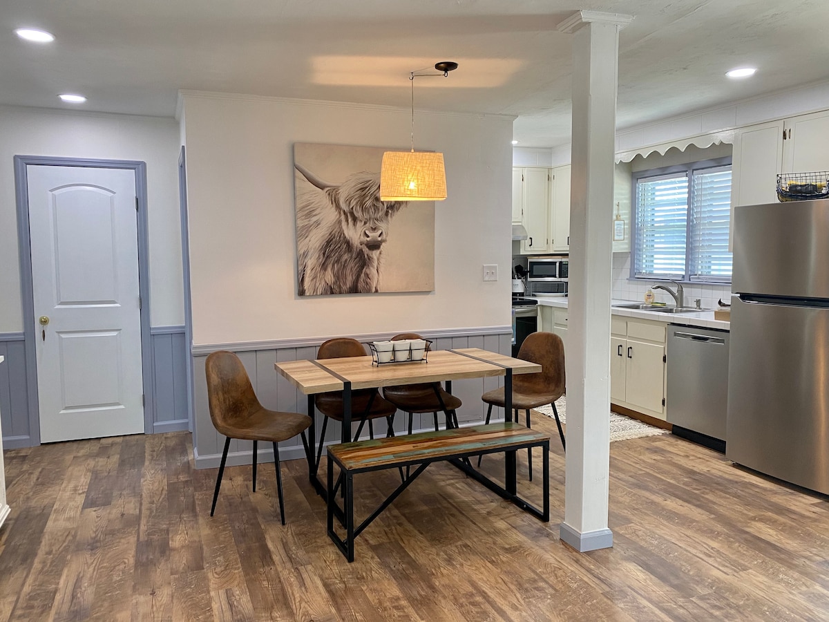 A dining area is presented, featuring a wooden table with bench seating surrounded by four chairs. A soft light fixture hangs above, illuminating a wall adorned with a large animal portrait. The kitchen area can be seen in the background, showcasing modern appliances.