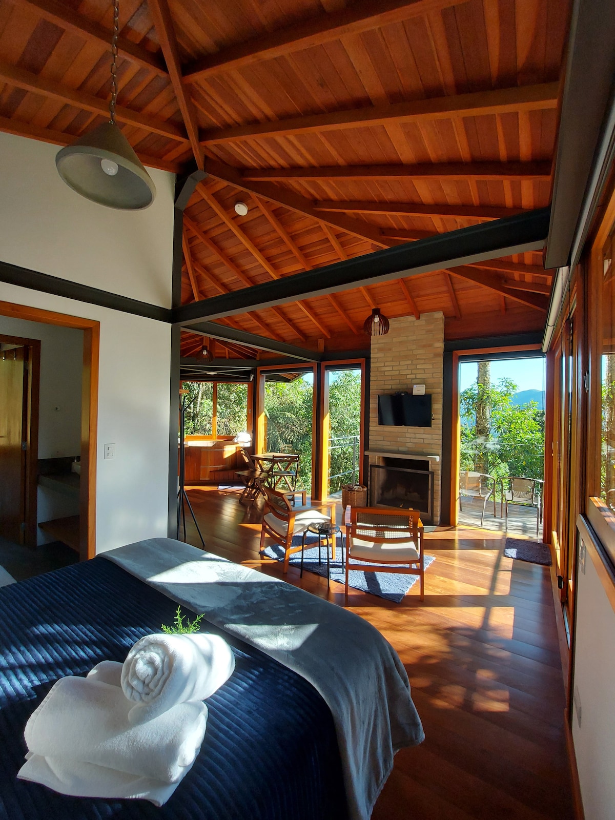 A spacious interior is presented, featuring a high wooden ceiling and large windows that offer views of the surrounding nature. A cozy seating area with chairs is positioned near a fireplace, while a comfortable bed with neatly arranged towels occupies the foreground.