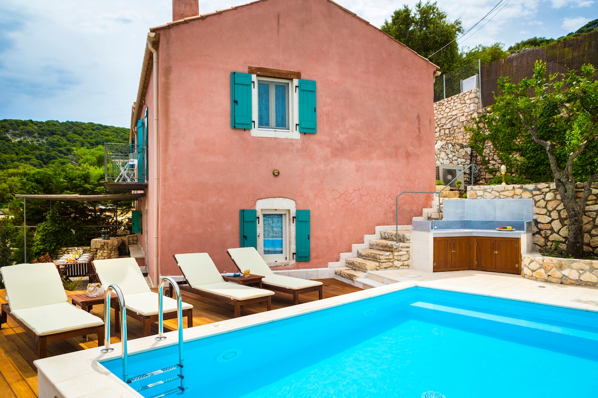 A charming pink villa is set against a backdrop of lush greenery. A private pool is surrounded by wooden decking and comfortable loungers. An outdoor kitchen area is visible, enhancing the inviting ambiance of the space. The villa's turquoise shutters add a colorful touch.