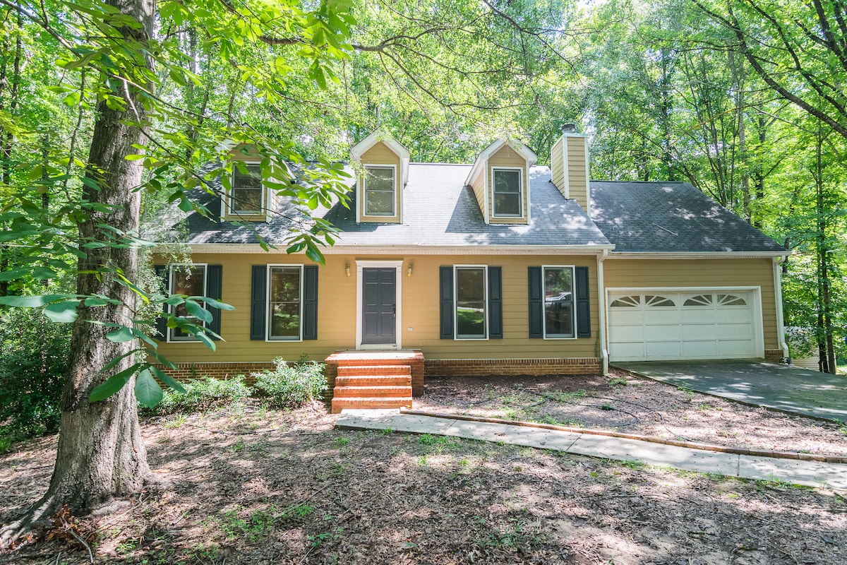 The home features a two-story exterior with a beige facade and classic black shutters. A spacious driveway leads to a two-car garage. Surrounding trees provide a wooded backdrop, enhancing the sense of privacy and tranquility in the front yard.