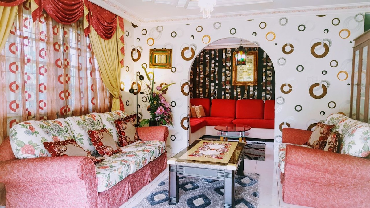 A vibrant living room is presented with floral-patterned sofas and a bright red sectional. Decorative curtains frame the windows while a coffee table occupies the center, adorned with a decorative tablecloth. A framed piece of art and potted flowers enhance the cheerful atmosphere.