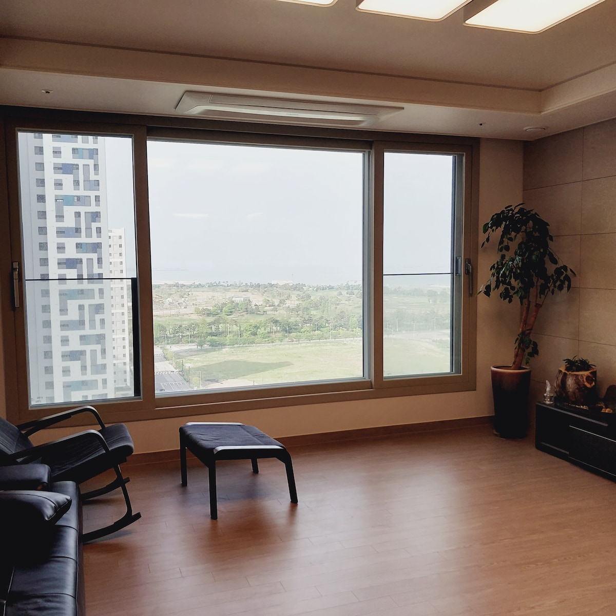 An inviting living area features large windows that offer a view of green spaces and distant water. A comfortable black chair is positioned nearby, while a potted plant adds a touch of nature to the room. Natural light brightens the wooden flooring.