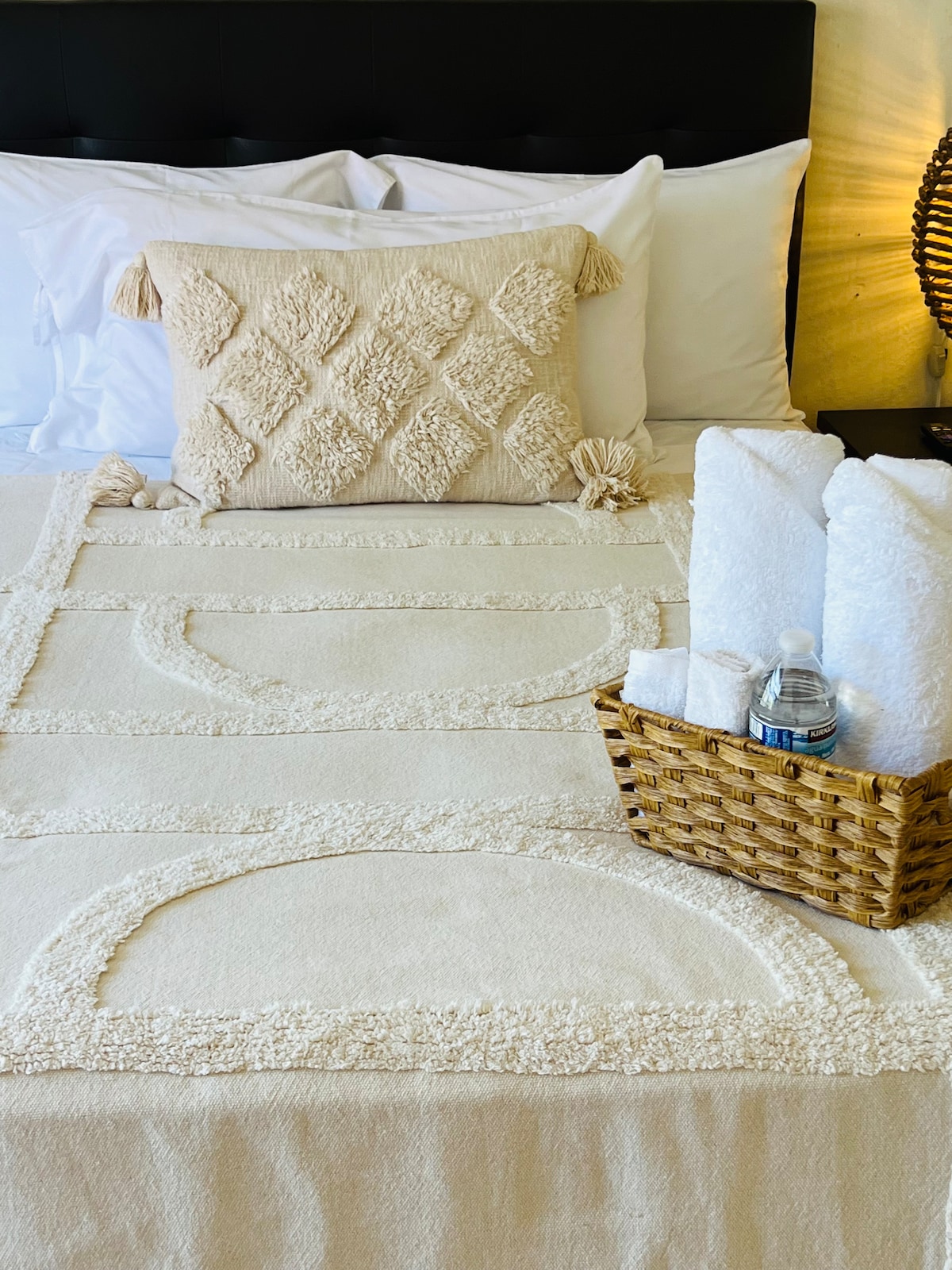 A neatly made bed features a soft cream-colored bedspread topped with decorative pillows. To the side, a woven basket holds bottled water and several neatly rolled white towels, providing cozy and convenient amenities for guests.