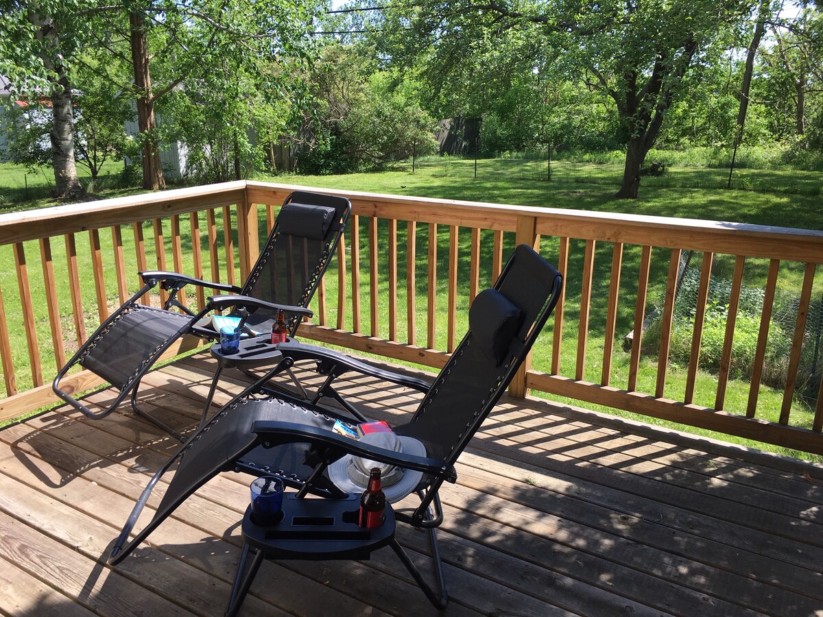 A deck is equipped with two reclining lounge chairs, positioned to offer comfort and relaxation. Sunlight filters through the surrounding greenery, casting gentle shadows on the wooden flooring. The spacious backyard is visible beyond the deck, featuring lush grass and scattered trees.
