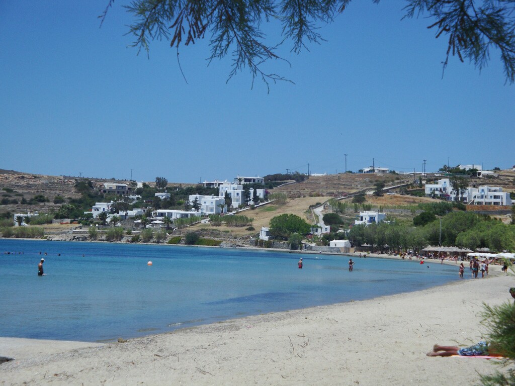 A serene sandy beach stretches along a clear blue coastline, with gentle waves lapping at the shore. White buildings are visible in the distance, nestled among green trees and rolling hills. People can be seen enjoying the water and sun, creating a lively yet relaxed atmosphere.