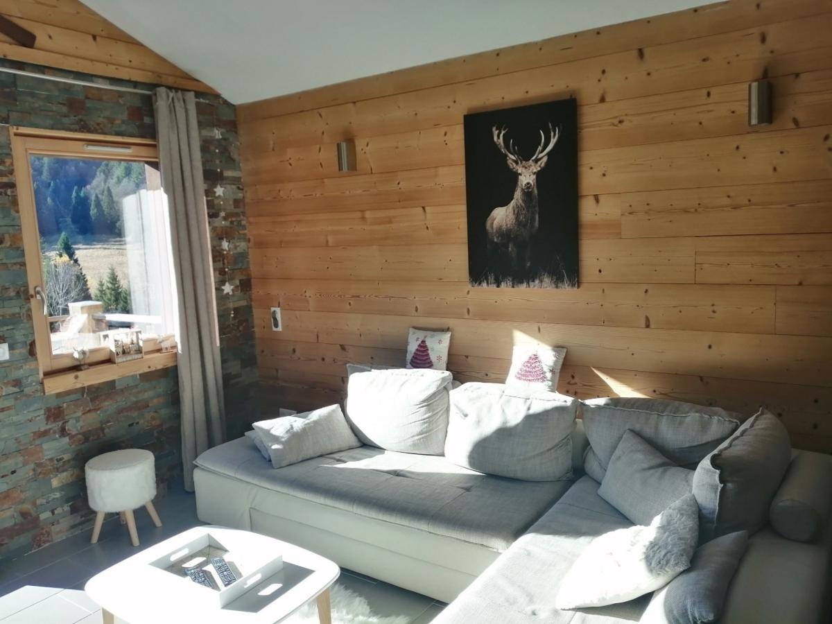 A light-filled living space features a spacious gray sofa against a wooden wall, complemented by a framed artwork of a deer. Nearby, a window offers views of the surrounding nature, while a small, round ottoman adds functionality.