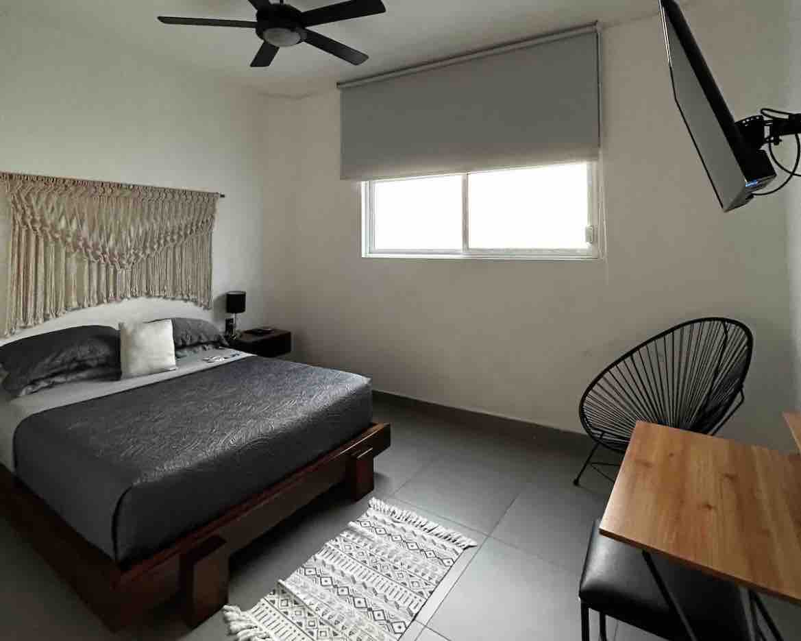 A comfortable sleeping area features a spacious bed with a dark grey coverlet. Natural light enters through a window covered by light grey blinds. A minimalist desk and chair are positioned beside the bed, while a television is mounted on the wall opposite the bed. 