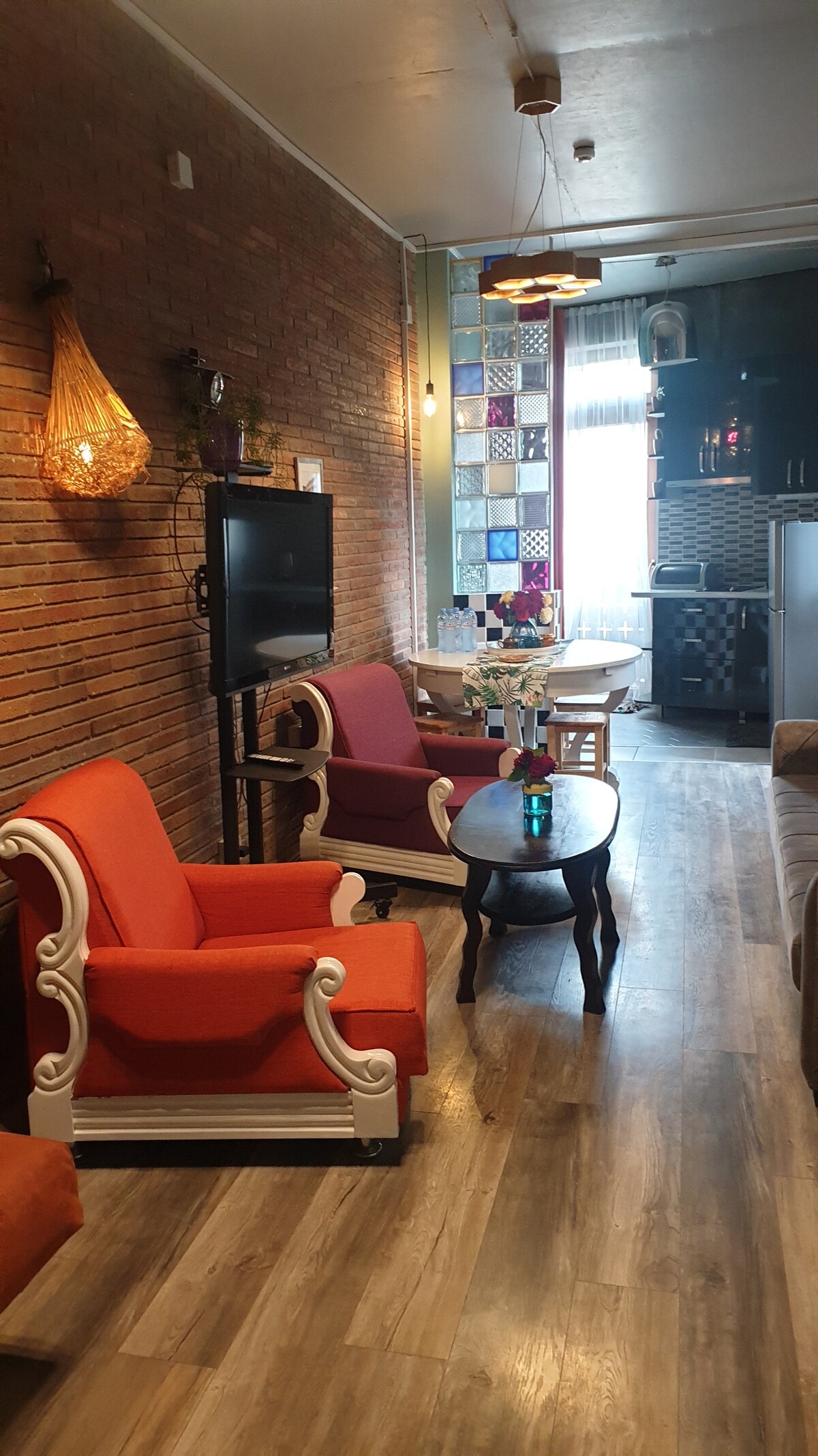 A cozy living area features two orange and purple chairs beside a small dark wooden table. A modest television stands against a brick wall, while a dining area with a table and chairs is visible in the background, illuminated by natural light from the window.