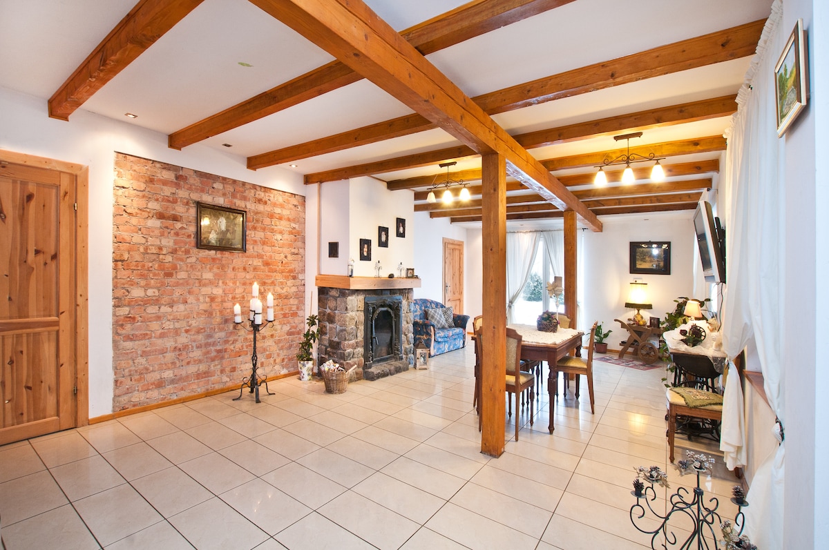 A spacious living area is highlighted by exposed wooden beams and a warm brick wall. A stone fireplace serves as a central feature, surrounded by cozy seating and a dining table. Large windows allow natural light to fill the room, enhancing the inviting ambiance.