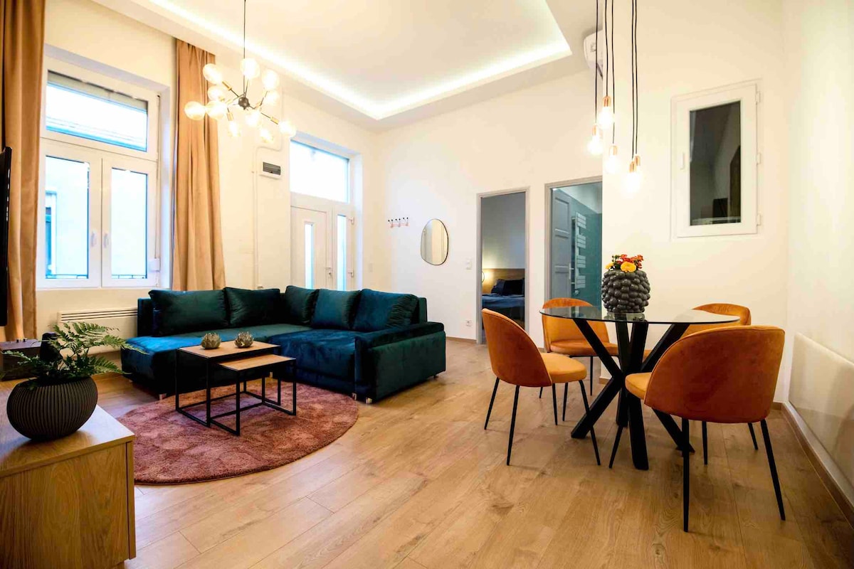 A modern living area features a plush green sofa, a coffee table, and a dining set with orange chairs. Natural light fills the space through multiple windows, complemented by elegant pendant lights. A round rug adds warmth, while a doorway leads to a bedroom.