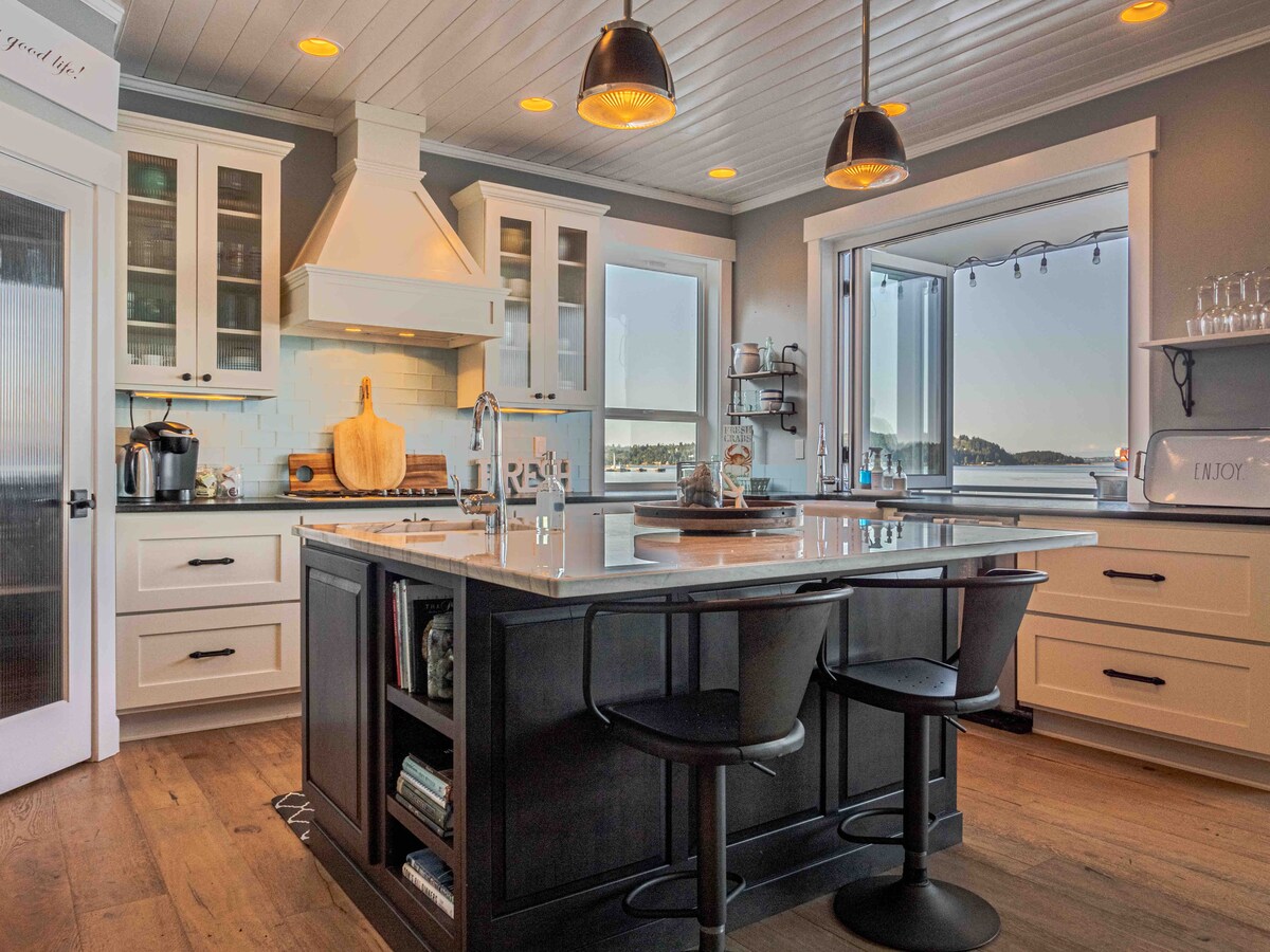 An open kitchen features modern cabinetry with glass doors, a large island with seating, and stylish pendant lighting. A panoramic window provides breathtaking views of the surroundings while allowing natural light to illuminate the space.