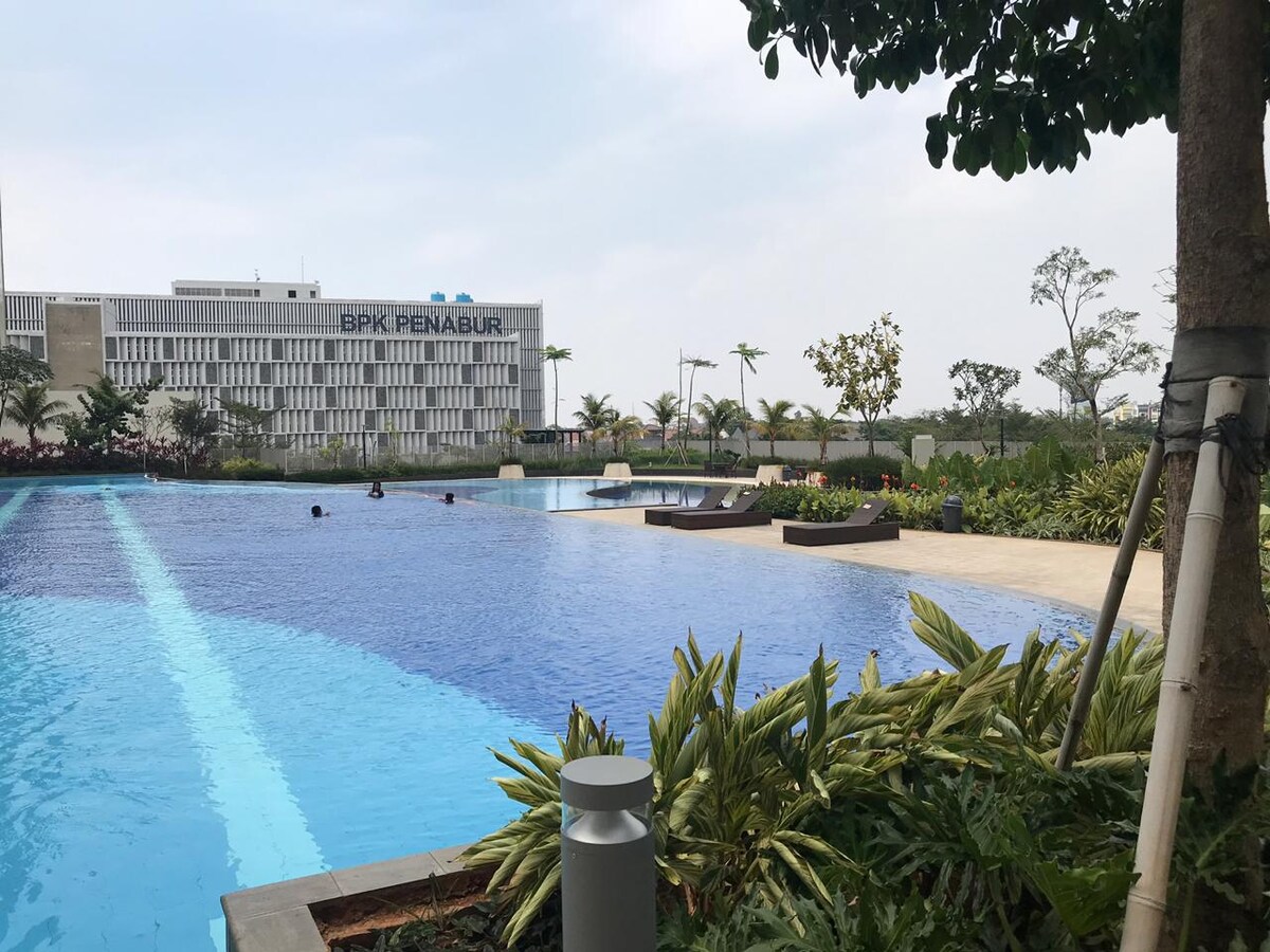 An expansive outdoor swimming pool is featured, surrounded by well-maintained greenery and landscaped areas. Lounge chairs are positioned along the poolside, providing a serene atmosphere. In the background, a modern building is visible, indicating proximity to local amenities.