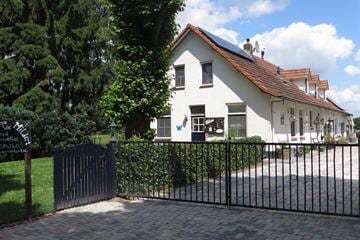The charming farmhouse features a welcoming entrance with a gated driveway. Lush greenery surrounds the property, and the building showcases a classic design with white walls and a traditional roof. Outdoor seating areas are visible, inviting guests to enjoy the peaceful surroundings.